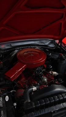An engine compartment of a vehicle with a predominantly red air filter cover and various mechanical components. The hood is open, revealing parts like hoses, wires, and metallic elements surrounding the engine block.