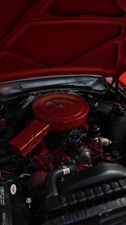 An engine compartment of a vehicle with a predominantly red air filter cover and various mechanical components. The hood is open, revealing parts like hoses, wires, and metallic elements surrounding the engine block.