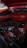 An engine bay of a classic car with a prominently displayed red air filter cover. The engine components are visible and include various hoses, wires, and a distributor. There is a mix of metal and rubber materials, and the car's exterior is painted in a matching red color.