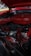 An engine bay of a classic car with a prominently displayed red air filter cover. The engine components are visible and include various hoses, wires, and a distributor. There is a mix of metal and rubber materials, and the car's exterior is painted in a matching red color.