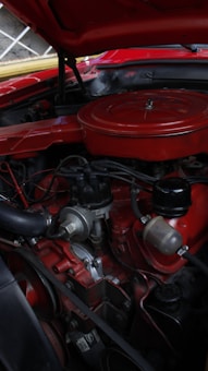 An engine bay of a classic car with a prominently displayed red air filter cover. The engine components are visible and include various hoses, wires, and a distributor. There is a mix of metal and rubber materials, and the car's exterior is painted in a matching red color.