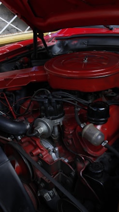 An engine bay of a classic car with a prominently displayed red air filter cover. The engine components are visible and include various hoses, wires, and a distributor. There is a mix of metal and rubber materials, and the car's exterior is painted in a matching red color.