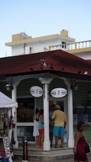 A small 7-Eleven store with a group of people standing outside, possibly tourists. The store has a green exterior and a red roof, with birds perched on top. There is a display of souvenirs and trinkets nearby, suggesting a tourist-friendly location. The background features white buildings with architectural details like railings.