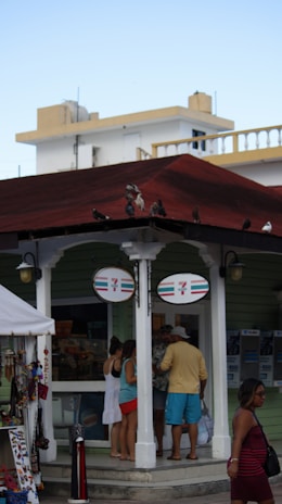 A small 7-Eleven store with a group of people standing outside, possibly tourists. The store has a green exterior and a red roof, with birds perched on top. There is a display of souvenirs and trinkets nearby, suggesting a tourist-friendly location. The background features white buildings with architectural details like railings.