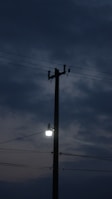 A tall utility pole stands against a backdrop of a dark, cloudy sky. A bright lamp is attached to the pole, casting a small area of light. Several electrical wires are visible extending from the pole.