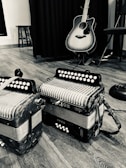 A black and white photo capturing two accordions placed on a wooden floor accompanied by an acoustic guitar leaned against a curtain. The setting suggests a musical environment, enhanced by a stool partially visible in the background.