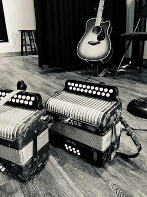 A black and white photo capturing two accordions placed on a wooden floor accompanied by an acoustic guitar leaned against a curtain. The setting suggests a musical environment, enhanced by a stool partially visible in the background.