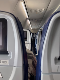 A view of an airplane cabin showcasing rows of passenger seats with personal entertainment screens. The overhead panel is visible with various air vents and light controls. One passenger is partially visible, seated in the aisle.