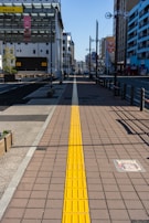 A long pedestrian walkway with a yellow tactile paving strip runs through the center, guiding travelers along the path. Buildings line both sides of the street, with banners hanging from lampposts. In the distance, a clear blue sky is visible, creating a bright and sunny urban environment.