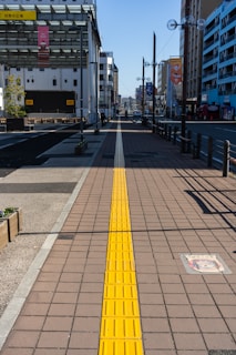 A long pedestrian walkway with a yellow tactile paving strip runs through the center, guiding travelers along the path. Buildings line both sides of the street, with banners hanging from lampposts. In the distance, a clear blue sky is visible, creating a bright and sunny urban environment.