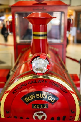 A close-up view of a miniature red locomotive with the words 'Kyushu Railway Company' and 'Bun Bun Go 2012' displayed on the front. The train has a shiny red finish with golden accents and a visible headlight in the center.