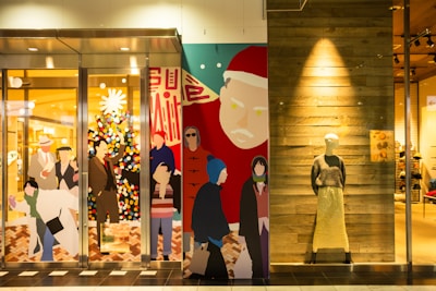 The image features a storefront with colorful, stylized graphics of people shopping and a decorated Christmas tree. A large depiction of Santa Claus is on one side. To the right, a mannequin wearing a sweater and long skirt is displayed against a wooden wall.