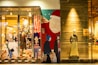 The image features a storefront with colorful, stylized graphics of people shopping and a decorated Christmas tree. A large depiction of Santa Claus is on one side. To the right, a mannequin wearing a sweater and long skirt is displayed against a wooden wall.