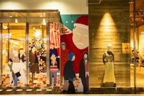 The image features a storefront with colorful, stylized graphics of people shopping and a decorated Christmas tree. A large depiction of Santa Claus is on one side. To the right, a mannequin wearing a sweater and long skirt is displayed against a wooden wall.