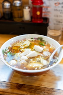 A bowl of soup with various ingredients including white tofu blocks, boiled eggs, and vegetables in a rich broth. The soup is served in a decorative bowl placed on a wooden table. In the background, there are condiment containers and a blurred menu.