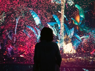 A silhouette of a person stands in front of a colorful, digital art display featuring lush digital foliage and an array of pink and blue particles swirling around a bright tree.