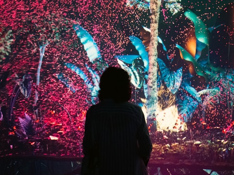 A silhouette of a person stands in front of a colorful, digital art display featuring lush digital foliage and an array of pink and blue particles swirling around a bright tree.