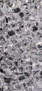 A densely packed cluster of clear ice cubes, mostly square in shape, with light reflecting off their surfaces creating a shiny appearance.