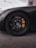 A close-up view of a black car wheel showcasing a detailed alloy rim with a prominent brake caliper. The wheel is on a textured asphalt surface, with part of the car body visible. The logo on the wheel hub indicates a luxury car brand. A palm tree and lamp post are faintly visible in the background against a cloudy sky.