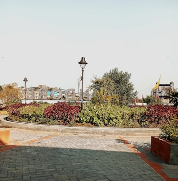 A well-maintained public garden area with paved pathways, vibrant red and green foliage, and street lamps. The background features a sizable sign structure with letters and partially visible urban buildings.