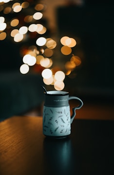 A ceramic mug decorated with botanical designs sits on a wooden surface. In the background, there is a soft bokeh effect with warm, circular lights, suggesting a cozy, ambient atmosphere.