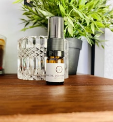 A small brown bottle labeled as massage oil or body wash is placed on a wooden surface. Behind it, there is a textured glass container and a black potted plant with green leaves. The setting appears to be a calm and natural environment with household or spa-like items.