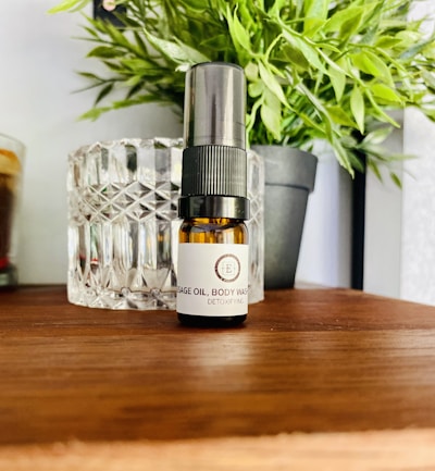 A small brown bottle labeled as massage oil or body wash is placed on a wooden surface. Behind it, there is a textured glass container and a black potted plant with green leaves. The setting appears to be a calm and natural environment with household or spa-like items.