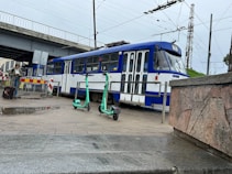 A blue and white tram is passing under a bridge on a paved street. Two electric scooters are parked nearby. A person is standing with a suitcase and looking at a mobile device. The tram tracks are visible, and overhead wires crisscross the sky. The scene appears to be urban with some greenery in the background.