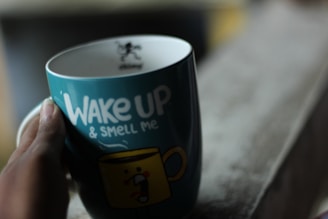 A hand holding a teal-colored mug with the phrase 'Wake Up & Smell Me' printed on it. The design includes a stylized, cartoonish yellow cup with a facial expression. The scene is softly lit, giving a cozy and intimate feel.