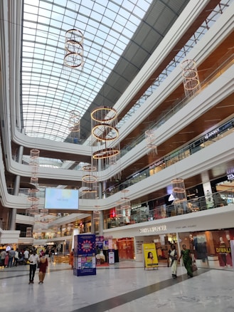 A modern shopping mall interior with multiple levels and an expansive glass ceiling. The architecture features clean lines and a spacious layout. Several floors of shops are visible, including brands like Marks & Spencer and Adidas. Pendant lights with circular designs hang from above, adding an elegant touch. People are strolling through the open space, and a promotional display for Diwali is prominent in the center.