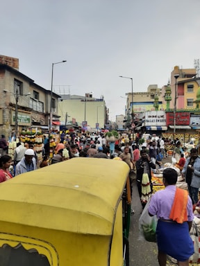 A bustling street market scene filled with numerous people walking and engaging in activities. The street is lined with colorful stalls selling flowers, clothing, and assorted goods. There is a yellow auto-rickshaw in the foreground. The buildings display vibrant signage with various shop names, and hanging garlands add to the lively atmosphere.