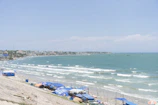 A coastal scene with a sandy beach stretching along the shoreline, dotted with small blue and white tents or umbrellas. The ocean's turquoise waves roll gently towards the shore, and a town with a cluster of buildings is visible in the distance along the coast. The sky is clear and blue, and the overall atmosphere is serene and summery.