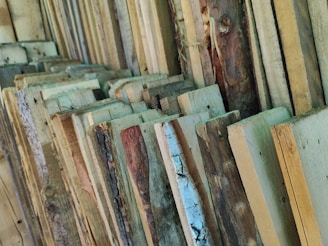 A collection of rough-cut wooden planks arranged in a vertical position, displaying various natural textures and shades of brown and beige. The wood grain and some knots are visible, conveying the raw and rustic nature of the material.