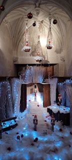 An interior space decorated for the holiday season with a winter theme. The scene includes several nutcracker dolls and artificial snow, creating a festive atmosphere. There are glowing blue lights and white branches, and hanging from the ceiling are decorative elements resembling large ornaments and light fixtures. The environment has a cozy, magical essence.