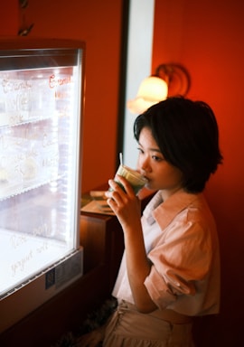A person is standing next to a refrigerator with a glass door, drinking from a green beverage cup. There is writing on the refrigerator door with various menu items. The setting has warm lighting, and there is a decorative lamp in the background.