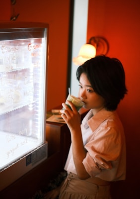 A person is standing next to a refrigerator with a glass door, drinking from a green beverage cup. There is writing on the refrigerator door with various menu items. The setting has warm lighting, and there is a decorative lamp in the background.