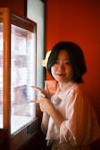 A person with short dark hair appears to be communicating expressively with their hands in front of a brightly lit display case. The warm red-orange hue of the walls and the soft lighting create a cozy ambiance.