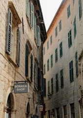 A narrow street lined with rustic stone buildings featuring green shutters on the windows. The architecture is traditional, with arched doorways and small shops, including one with a 'Bonaca Shop' sign. The image captures a sense of historic charm, enhanced by the aged stonework and the quietness of the alley.