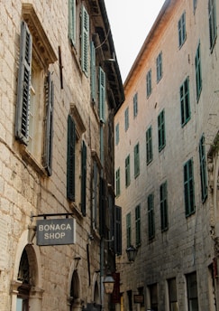 A narrow street lined with rustic stone buildings featuring green shutters on the windows. The architecture is traditional, with arched doorways and small shops, including one with a 'Bonaca Shop' sign. The image captures a sense of historic charm, enhanced by the aged stonework and the quietness of the alley.