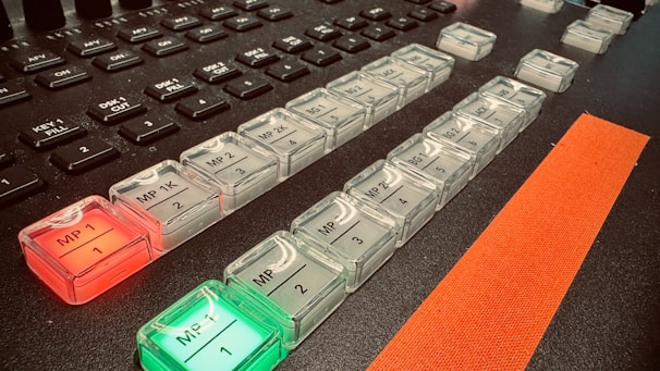 A close-up of a control panel featuring rows of illuminated buttons with labels such as 'MP 1K', 'MP 2', and others. The buttons are backlit with red and green lights. There are additional unlit keys above the buttons, which appear darker. A prominent orange strip runs alongside the buttons, adding a contrasting element.