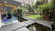 A modern outdoor space features a glass-walled building with seating inside, adjacent to a landscaped garden with lush greenery. A small water feature runs alongside a stone pathway that leads into the garden. The green space includes several trees and plants, with tables and umbrellas visible in the background. The area appears well-maintained and designed for relaxation.