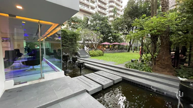 A modern outdoor space features a glass-walled building with seating inside, adjacent to a landscaped garden with lush greenery. A small water feature runs alongside a stone pathway that leads into the garden. The green space includes several trees and plants, with tables and umbrellas visible in the background. The area appears well-maintained and designed for relaxation.