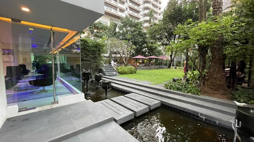 A modern outdoor space features a glass-walled building with seating inside, adjacent to a landscaped garden with lush greenery. A small water feature runs alongside a stone pathway that leads into the garden. The green space includes several trees and plants, with tables and umbrellas visible in the background. The area appears well-maintained and designed for relaxation.