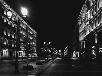 Black-and-white photo of an empty city street at night with deep shadows and glowing street lamps.