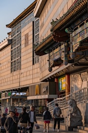 A street scene with people walking near a building that features both traditional Asian architectural elements and modern business signs, including a McDonald's logo. The structure showcases ornate, colorful decorations typical of Eastern design, juxtaposed with contemporary commercial signage. A statue resembling a stone lion is visible, adding a historical touch.