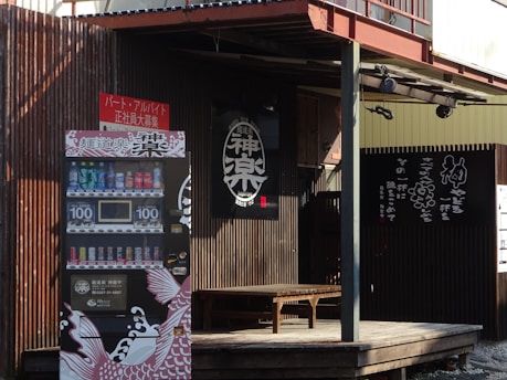 The scene features a rustic wooden facade of a building with a black sign displaying Japanese characters. A vending machine with various drinks, priced at 100 yen, is located at the front. The design of the vending machine includes artistic elements like stylized fish graphics. A wooden bench is situated on a small deck near the entrance.