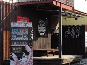 The scene features a rustic wooden facade of a building with a black sign displaying Japanese characters. A vending machine with various drinks, priced at 100 yen, is located at the front. The design of the vending machine includes artistic elements like stylized fish graphics. A wooden bench is situated on a small deck near the entrance.