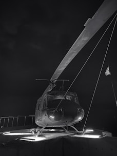 A helicopter is stationed on a landing pad at night. The scene is illuminated by artificial lighting, casting shadows and highlighting the helicopter's structure and blades against a dark sky.