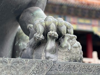 A detailed sculpture of a mythical creature, possibly a lion, is depicted with intricate carvings and patterns. It has a spherical object under one paw and exhibits traditional artistic motifs. The background is slightly out of focus, featuring part of a building with ornate architectural details.