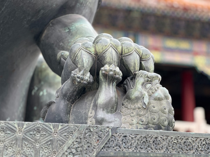 A detailed sculpture of a mythical creature, possibly a lion, is depicted with intricate carvings and patterns. It has a spherical object under one paw and exhibits traditional artistic motifs. The background is slightly out of focus, featuring part of a building with ornate architectural details.